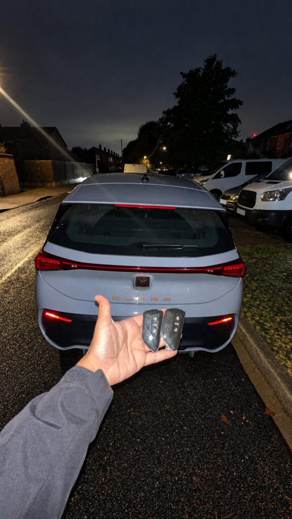 Two Cupra key fobs held in front of the rear of a 2023 electric car at night.